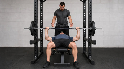 A lifter presses a loaded barbell off the chest on a flat bench while a spotter stands ready at the head of the bar.