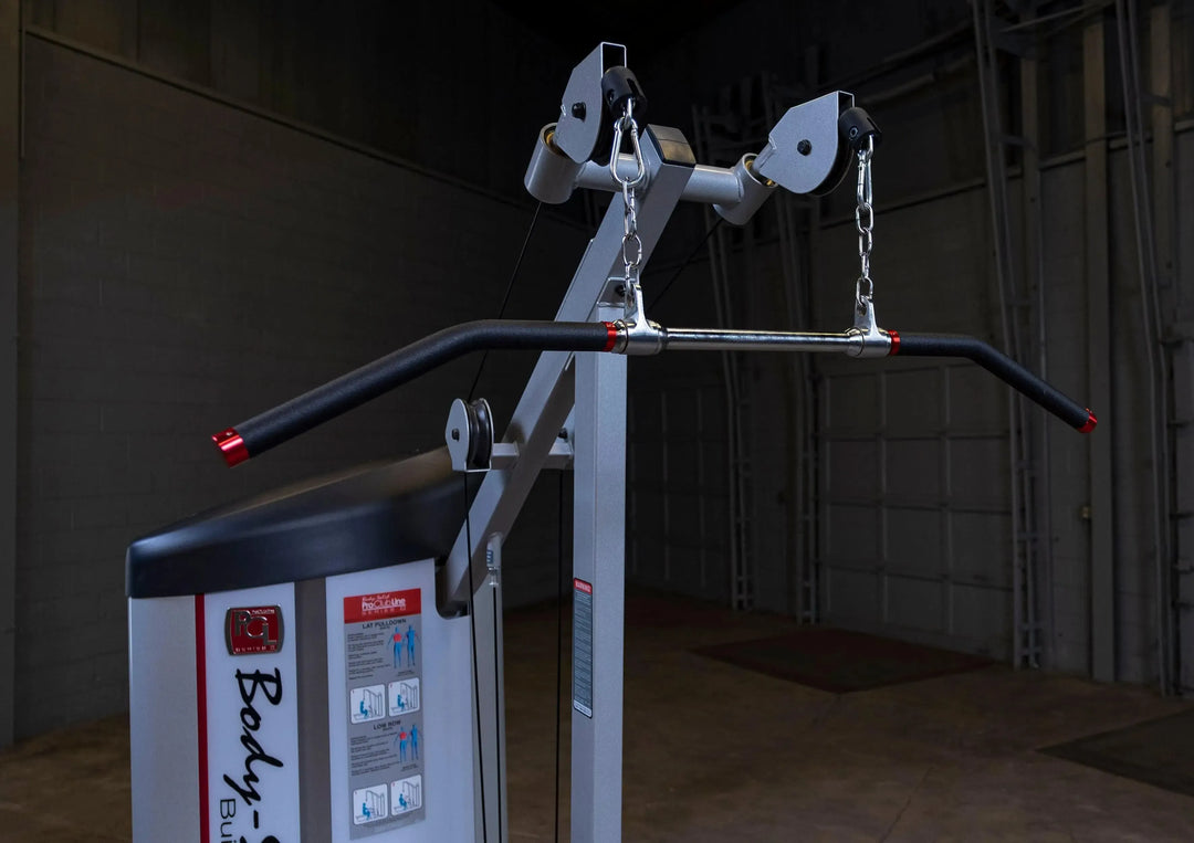 In a dimly lit gym with grey walls and flooring, a Body-Solid Pro Clubline Lat Pulldown and Row Machine (S2LAT) awaits. The pull-down attachment includes two chains and a curved handlebar, while workout instructions on the side cater to strength training enthusiasts.