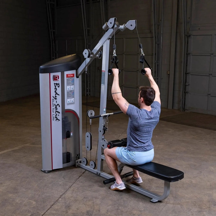In a dimly lit gym, someone in a gray shirt and blue shorts does strength training on the Body-Solid Pro Clubline Lat Pulldown and Row Machine (S2LAT), gripping the overhead bar while seated on a black bench.