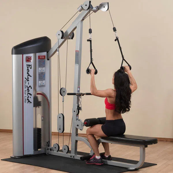 A person sits on a Body-Solid Pro Clubline Lat Pulldown and Row Machine (S2LAT), wearing a red sports bra and black shorts. The machines tall, rectangular frame with cables and pulleys underscores their dedication to strength training in a neutral setting.