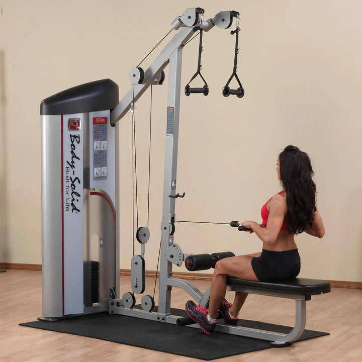 A person wearing a red sports bra and black shorts uses the Body-Solid Pro Clubline Lat Pulldown and Row Machine (S2LAT) on a black mat in a gym with beige walls and wooden flooring, focusing on strength training to build endurance and power.