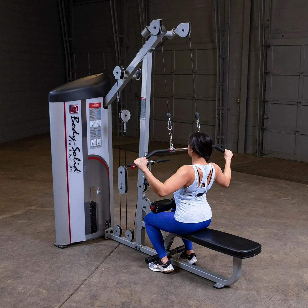 Wearing a white tank top and blue pants, a person uses a Body-Solid Pro Clubline Lat Pulldown and Row Machine (S2LAT) for strength training at the gym. They face the machine, gripping the bar with both hands on a concrete floor, with a garage door behind them.