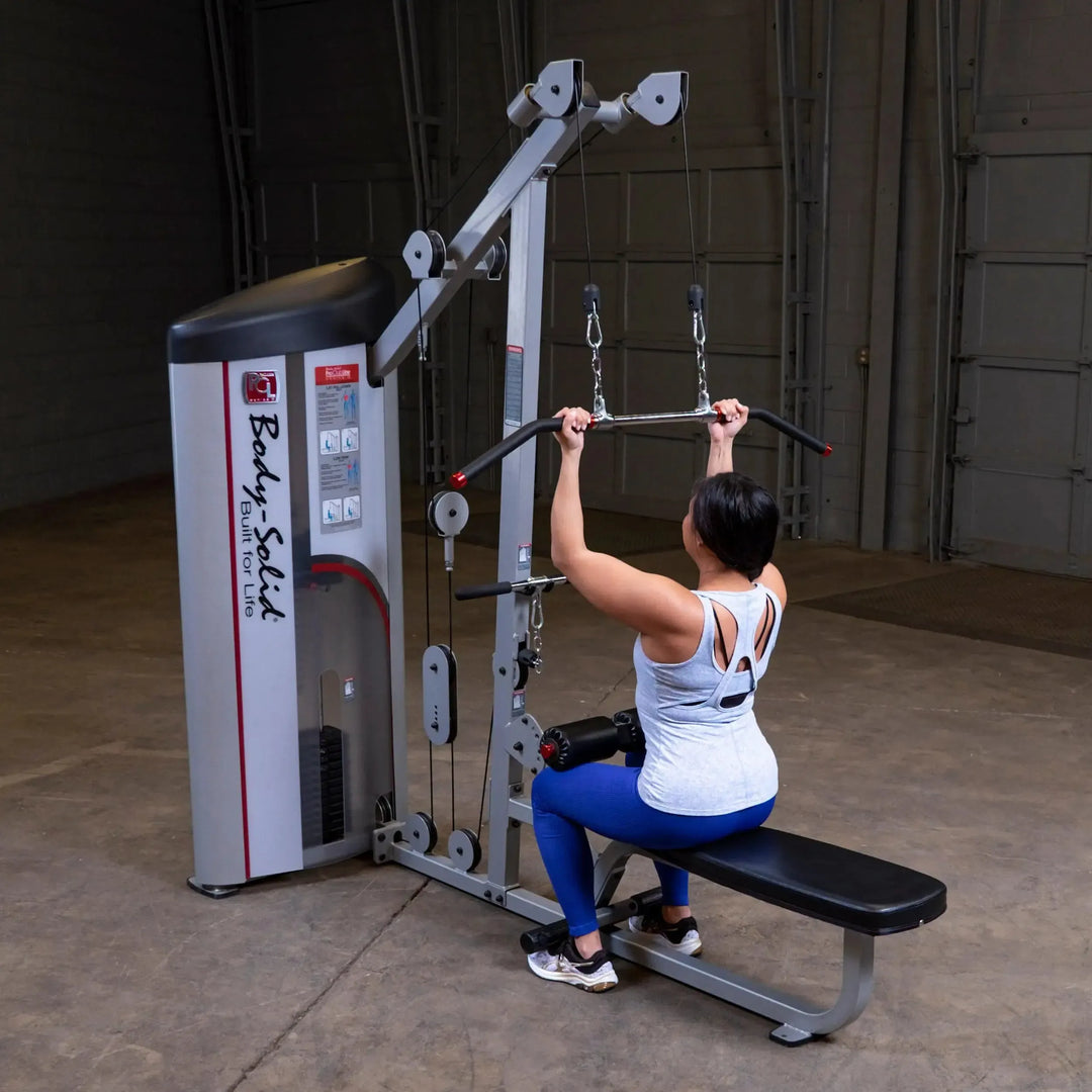 Wearing athletic clothing, a person uses the Body-Solid Pro Clubline Lat Pulldown and Row Machine (S2LAT) in a gym, pulling down a bar overhead while seated on a padded bench. The backdrop shows a spacious industrial setting perfect for strength training.
