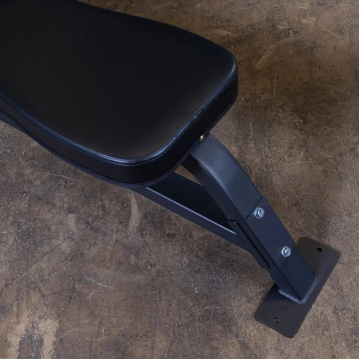 The Body-Solid Pro Clubline Olympic Flat Bench (SOFB250) rests on a textured concrete floor, showcasing its seat and sturdy metal base. Located in the upper left corner, it is securely bolted, making it a reliable addition to any fitness facility.