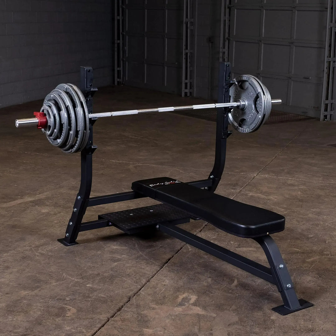 The Body-Solid Pro Clubline Olympic Flat Bench (SOFB250) is set up with a barbell and weights on a black bench in a gym. The concrete floor complements the gray wall and large door, showcasing this professional-grade equipment from Body-Solid Pro Clubline.