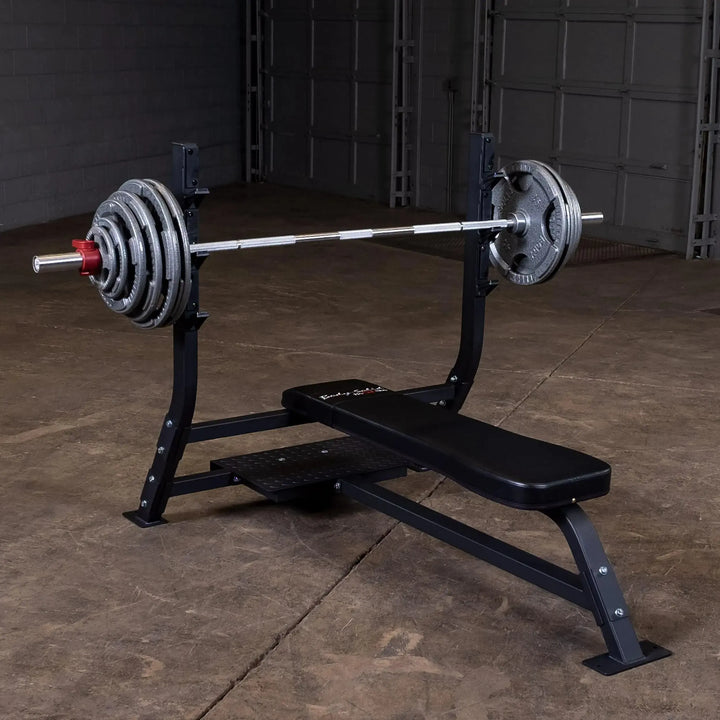 The Body-Solid Pro Clubline Olympic Flat Bench (SOFB250) is set up with a barbell and weights on a black bench in a gym. The concrete floor complements the gray wall and large door, showcasing this professional-grade equipment from Body-Solid Pro Clubline.