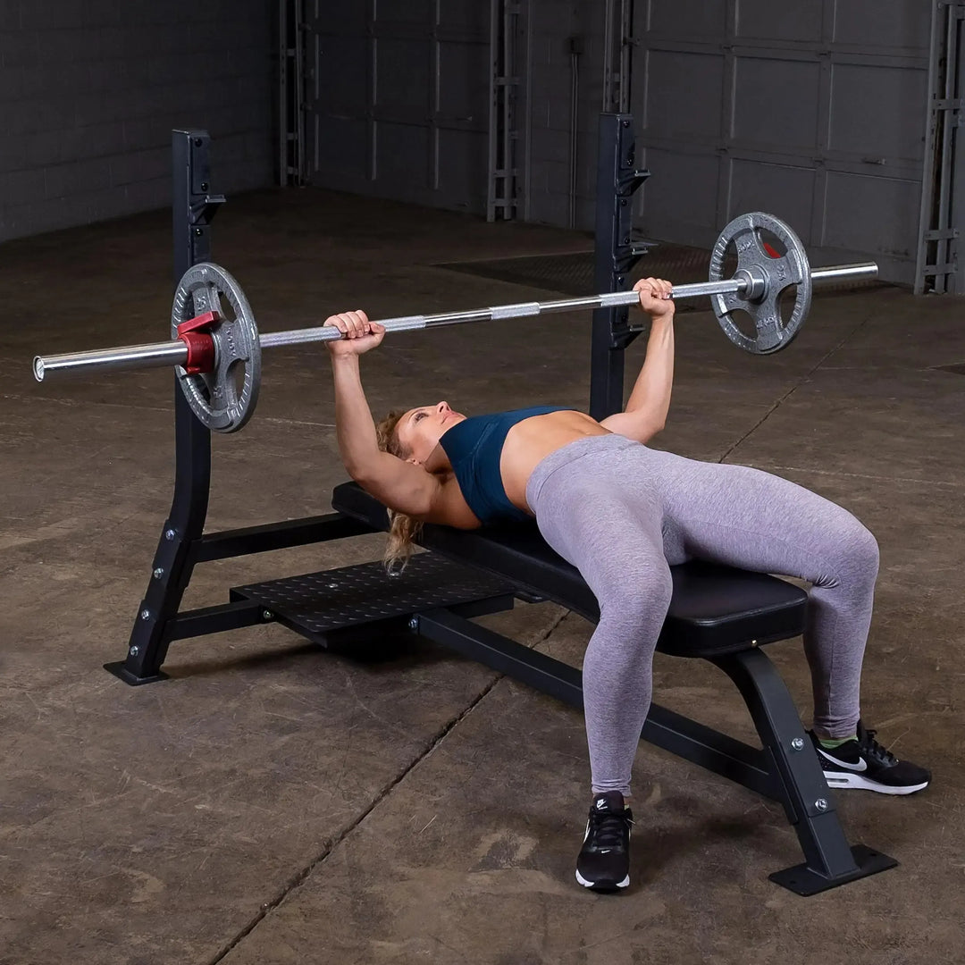 Wearing athletic gear, a person lies on a Body-Solid Pro Clubline Olympic Flat Bench (SOFB250) in a spacious gym, pressing a barbell with weights. They grip the barbell firmly with both hands, showcasing classic bench press form amid the fitness facilitys concrete floors and walls.