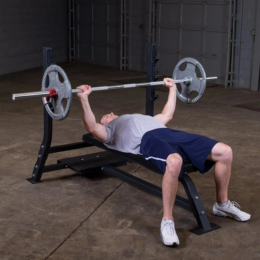 A person in a gray shirt and blue shorts performs a bench press on the Body-Solid Pro Clubline Olympic Flat Bench (SOFB250) in their garage gym, lifting a barbell with weighted plates as found in any fitness facility.