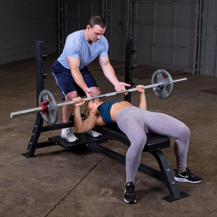 A woman in a blue sports bra and gray leggings skillfully bench presses on the Body-Solid Pro Clubline Olympic Flat Bench (SOFB250), with a man in a light blue shirt and navy shorts spotting her. They are surrounded by gym equipment on concrete floors in a large fitness facility with an open garage door.