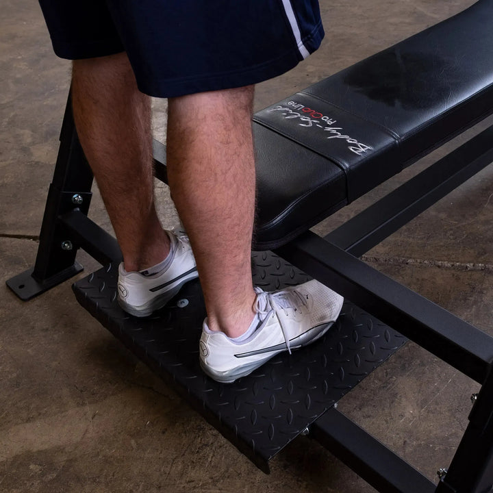 Close-up of a person wearing white sneakers and black shorts, heels raised on a step platform by the Body-Solid Pro Clubline Olympic Flat Bench (SOFB250). Only the legs are visible, highlighting this high-quality gym equipment. The concrete floor adds an industrial vibe to the scene.