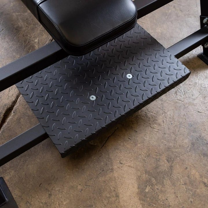 The close-up shows a black diamond-patterned metal footplate on a concrete floor, part of gym equipment beneath the padded Body-Solid Pro Clubline Olympic Flat Bench (SOFB250).