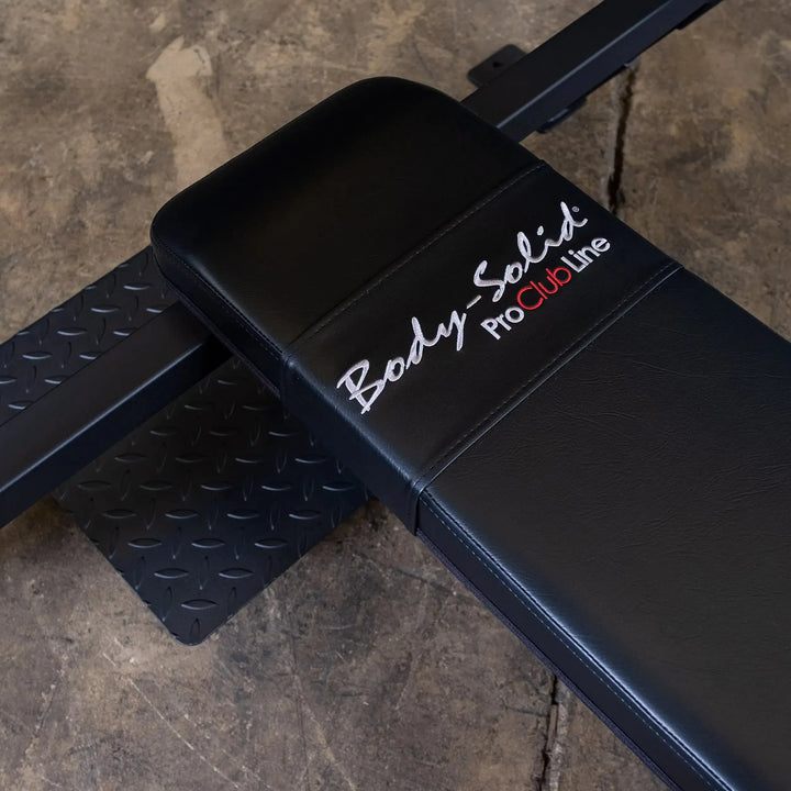 A close-up of the black Body-Solid Pro Clubline Olympic Flat Bench (SOFB250) features striking white and red branding. It rests on a textured metal floor, enhancing the gritty, industrial vibe of the fitness facilitys gym equipment area.