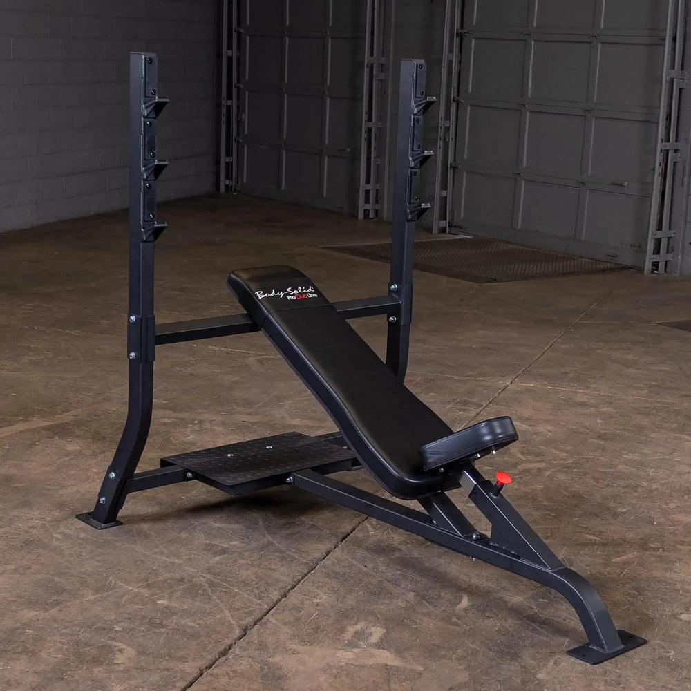 A Body-Solid Pro Clubline Olympic Incline Bench (SOIB250) with a barbell rack is in an empty room with concrete flooring and gray walls. This black, adjustable bench includes a stability footplate, ideal for upper body workouts and perfect for commercial fitness use.