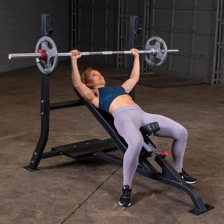 A woman in workout attire confidently uses a Body-Solid Pro Clubline Olympic Incline Bench (SOIB250) for a barbell press at the gym, focusing on sculpting her upper body with feet grounded and hands gripping the barbell.