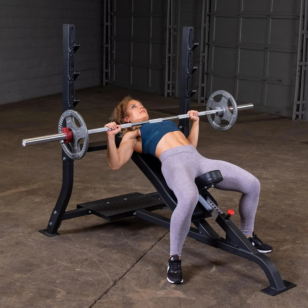In a gym with concrete walls and a metal garage-style door, someone in a blue sports top and gray leggings is focused on their upper body workout. They expertly lift heavy weights on the Body-Solid Pro Clubline Olympic Incline Bench (SOIB250), showcasing their dedication to fitness.