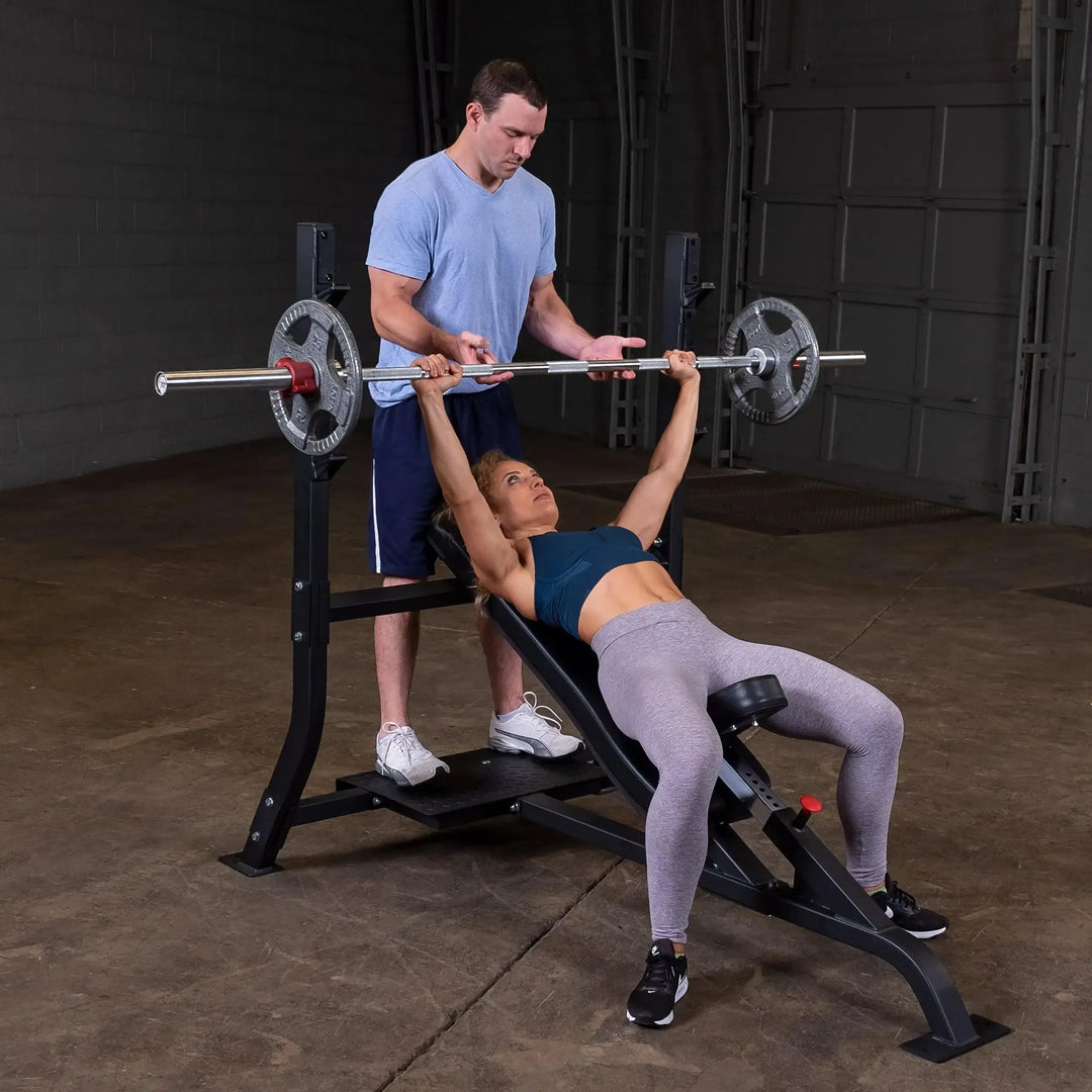 A person in a blue shirt observes someone adeptly using the Body-Solid Pro Clubline Olympic Incline Bench (SOIB250) in the gym. The focused lifter, wearing a teal sports bra and gray leggings, is amidst commercial fitness equipment surrounded by concrete floors and industrial walls.