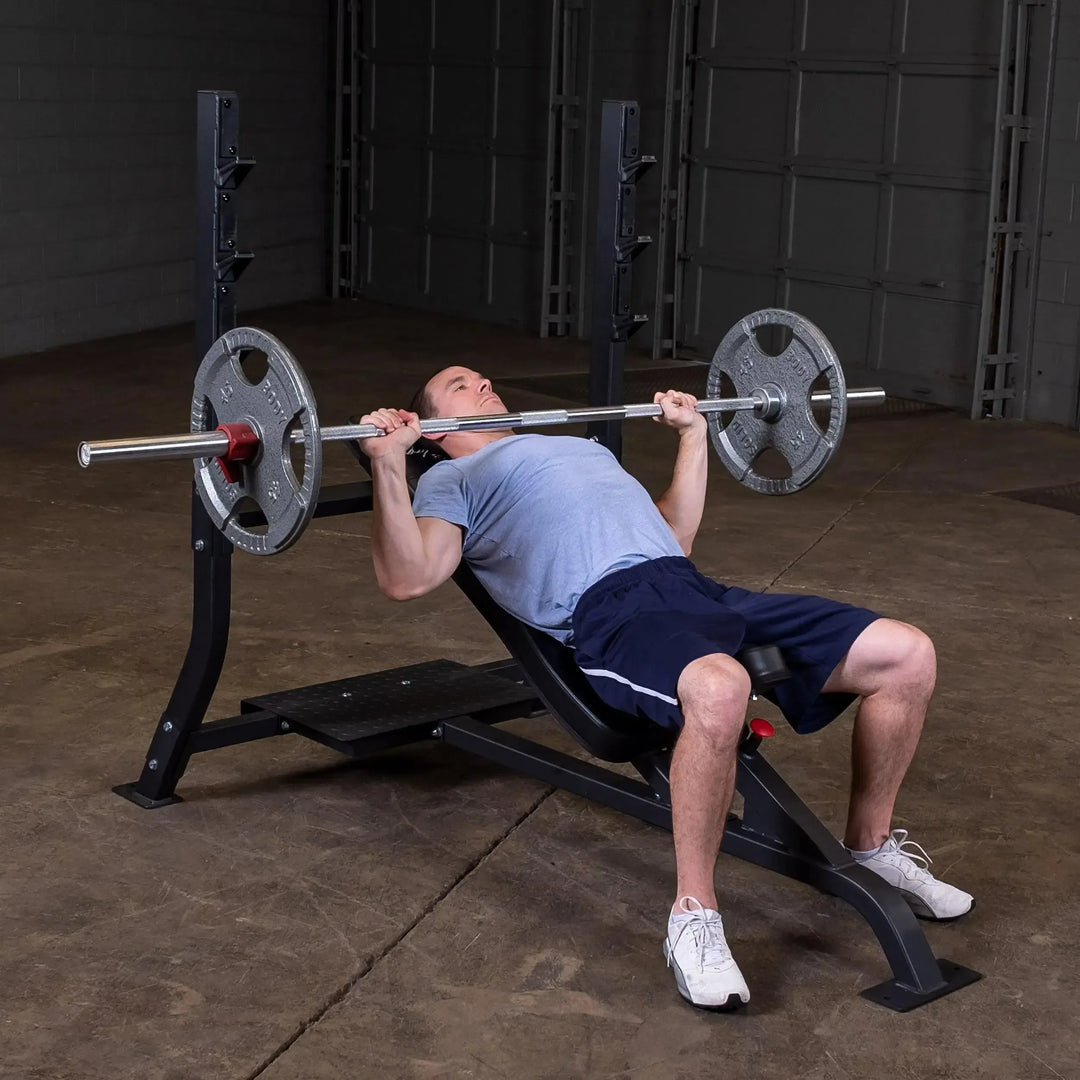 A person in a blue t-shirt and navy shorts does an upper body workout on the Body-Solid Pro Clubline Olympic Incline Bench (SOIB250) at the gym. Theyre lifting a barbell with weight plates in a power rack, surrounded by concrete floors and garage doors showcasing commercial fitness equipment.