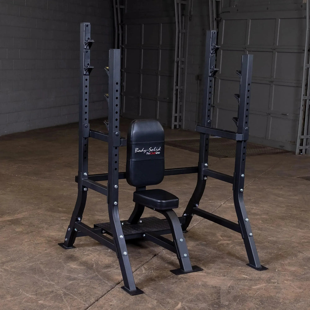 A Body-Solid Pro Clubline Olympic Shoulder Press Bench (SOSB250), featuring a heavy-duty steel mainframe, sits in an empty garage with concrete floors and a white brick wall. The equipment is perfect for strength training with its adjustable bench and barbell rack with safety stops.