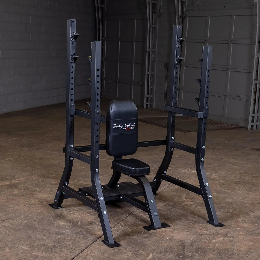 A Body-Solid Pro Clubline Olympic Shoulder Press Bench (SOSB250), featuring a heavy-duty steel mainframe, sits in an empty garage with concrete floors and a white brick wall. The equipment is perfect for strength training with its adjustable bench and barbell rack with safety stops.