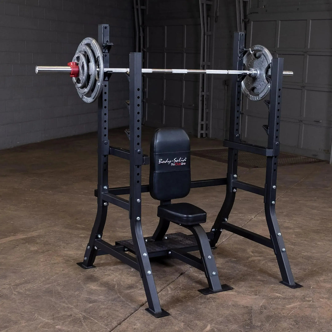 The Body-Solid Pro Clubline Olympic Shoulder Press Bench (SOSB250), made of heavy-duty steel, features a barbell, weight plates, adjustable safety stops, and a sleek black design. It sits on a brown concrete floor in a garage setting with a large closed door behind it.