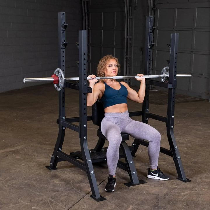 A curly-haired individual wearing a blue sports bra and gray leggings uses the Body-Solid Pro Clubline Olympic Shoulder Press Bench (SOSB250) with adjustable safety stops. Focused, they lift a barbell with weight plates in the gym.