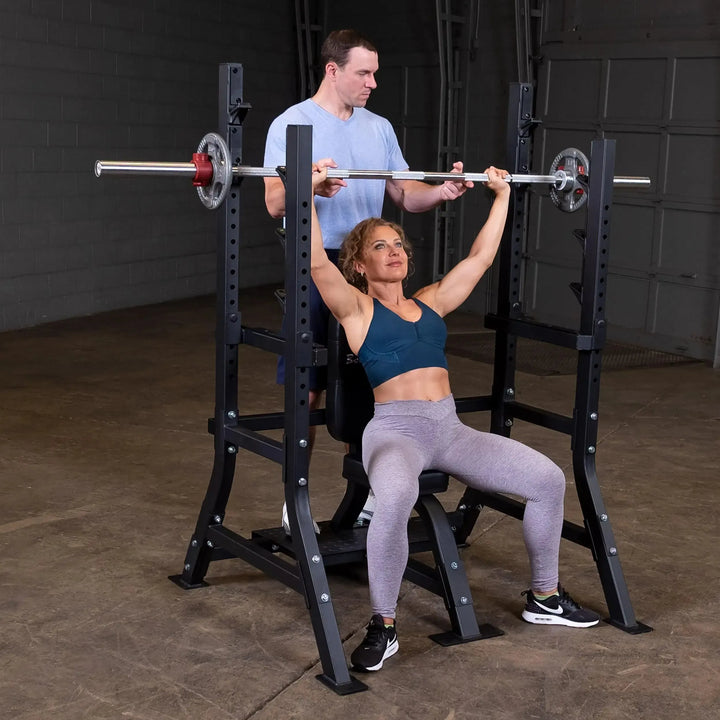 In a dimly-lit gym, a woman in teal sports bra and gray leggings lifts a barbell on the Body-Solid Pro Clubline Olympic Shoulder Press Bench (SOSB250) with adjustable safety stops as a man in a gray t-shirt spots her.