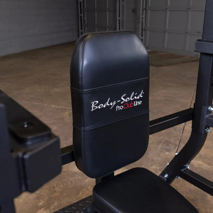 A black leather Body-Solid Pro Clubline Olympic Shoulder Press Bench (SOSB250) features the brand name on the backrest. In a gym with a concrete floor and garage door, its heavy-duty steel mainframe ensures durability.