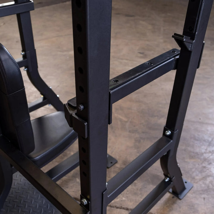 Close-up of the Body-Solid Pro Clubline Olympic Shoulder Press Bench (SOSB250) on a gyms concrete floor, showcasing a robust black metal frame with adjustable brackets and bolts. A padded seat partially visible on the left highlights the heavy-duty steel mainframe.