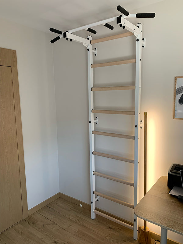 The BenchK Home Swedish Ladder w/ Pull-Up Bar (721) elegantly enhances a white wall, harmonizing with the rooms light wood flooring. Nearby, a desk and framed picture complete this chic home gym ambiance.