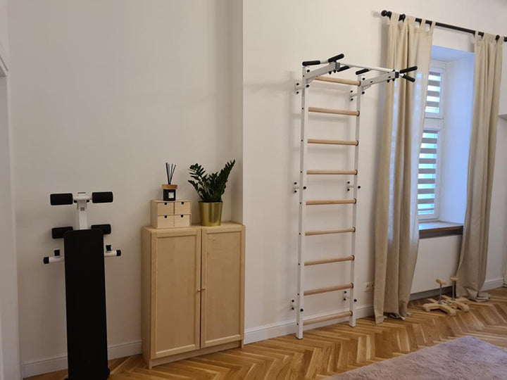The home gym area includes the BenchK Home Swedish Ladder with a wall-mounted pull-up bar and an adjustable black exercise bench next to a light wooden cabinet. A potted plant and decorative items adorn the cabinet, while cream curtains frame the window for a serene vibe.