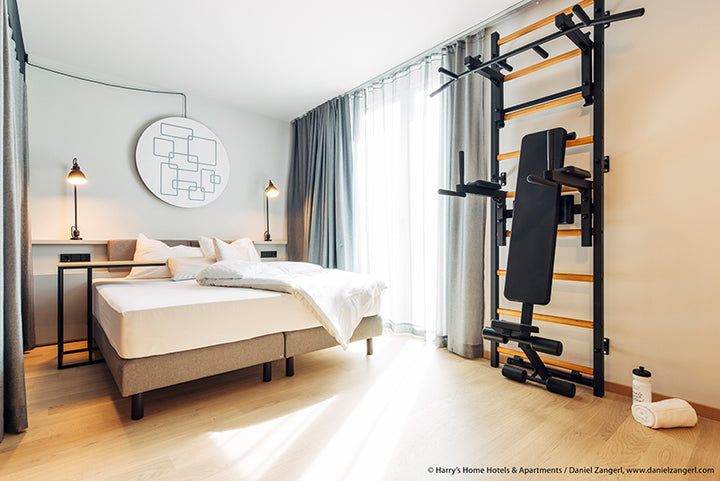 A modern bedroom features a neatly made double bed with white linens flanked by two bedside tables and lamps. Unique wall decoration adorns the area above the bed. Natural light flows through large curtained windows, while a BenchK Pro Gym Wall Bars w/ Bench, Dip & Flex Pull-Up Bar (733) stands on one side.