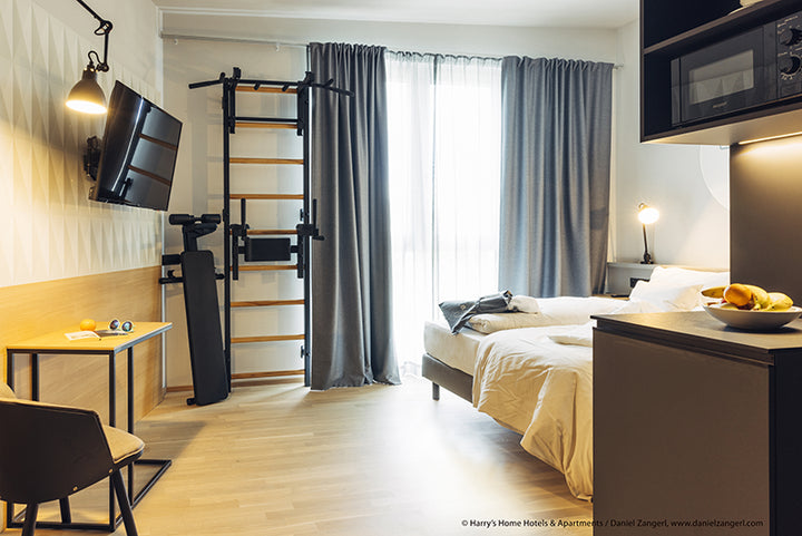 A modern hotel room features a bed, desk, chair, and wall-mounted TV. The space includes BenchKs Pro Gym Wall Bars with a bench, dip & flex pull-up bar (733) attached to the wall. Floor-to-ceiling curtains adorn the window, complemented by wooden flooring and minimalist decor.