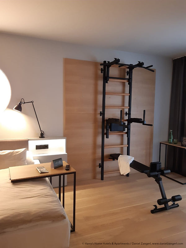 A modern hotel room showcases a bed with white linens, a desk lamp, and a nightstand with phone and alarm clock. BenchK Pro Gym Wall Bars w/ Bench and Dip & Flex Pull-Up Bar (733) serve as multifunctional wall equipment, with an exercise bike nearby. A window with curtains is on the right.