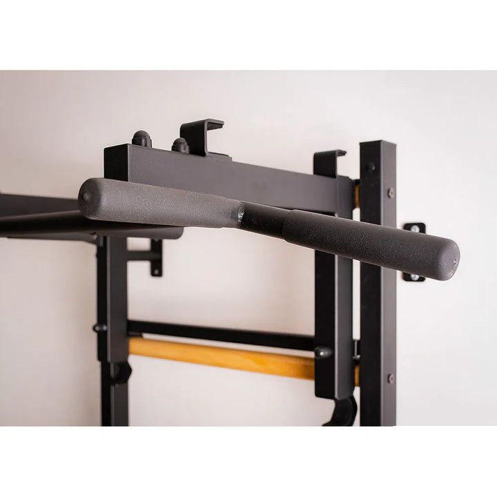 A close-up of the BenchK Pro Gym Wall Bars w/ Bench, Dip & Flex Pull-Up Bar (733) against a white wall shows its black metal bar with foam padding and sturdy power rack with vertical and horizontal supports, highlighting its versatile multifunctional design.