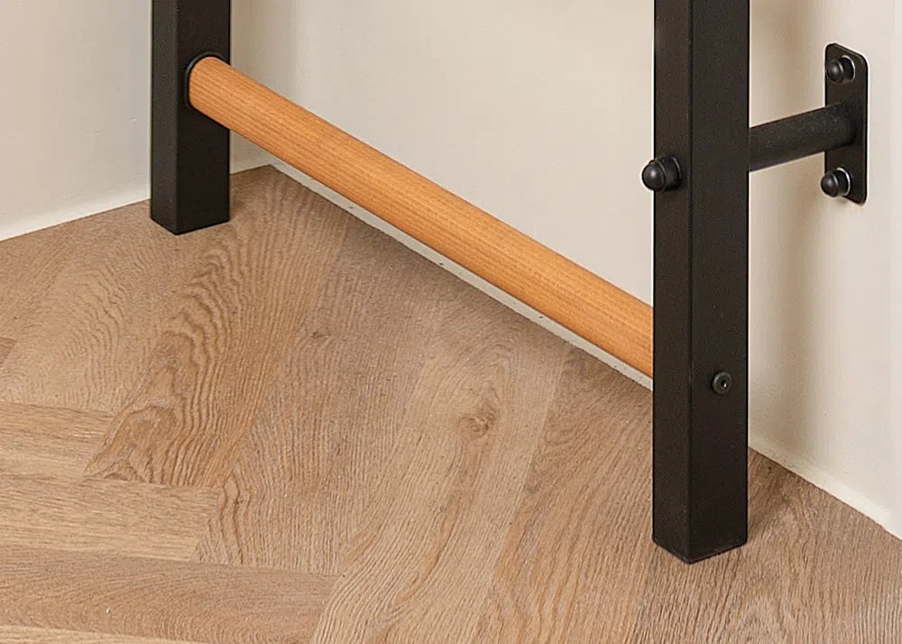 The BenchK Pro Gym Wall Bars with Bench, Dip & Flex Pull-Up Bar (733) features a wooden ballet barre on a black wall-mounted frame, securely fastened with screws against a light background and herringbone-patterned floor.