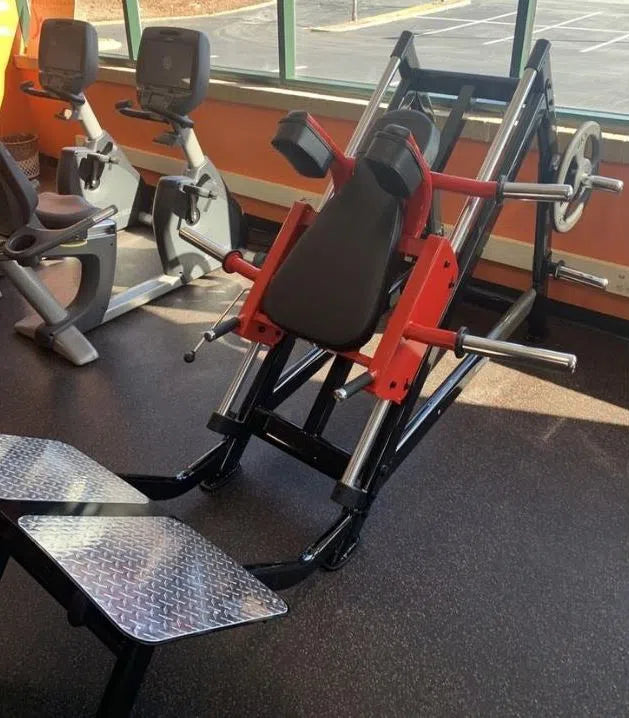 A SteelFlex Hack Squat Machine (PLHP), ideal for a lower body workout, sits next to two stationary bikes near a large window. The squat machine has red and black padding with metallic footplates, while the bikes feature digital screens and adjustable seats, typical of commercial fitness centers.