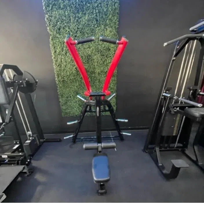 A Steelflex PLLA Lateral Pull Down Machine with red handles stands against a black wall, featuring a green textured panel. Its surrounded by other gym equipment on the dark floor and is essential in commercial gyms and fitness centers.