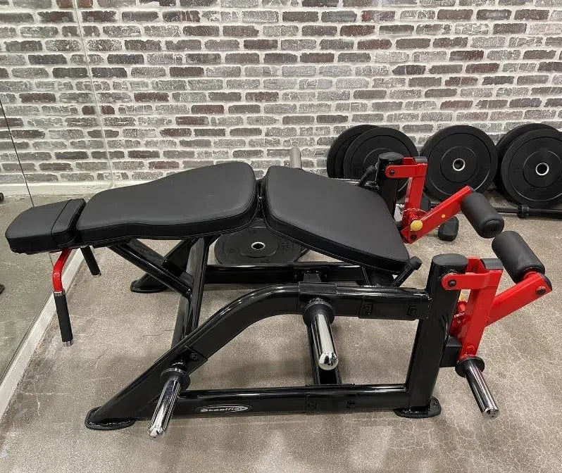 A Steelflex PLLC Hamstring Machine with black padding, red arm supports, and a sturdy black frame rests against a brick wall in the gym. Nearby, several black weight plates are neatly stacked on the floor, highlighting its commercial gym-quality equipment standards.