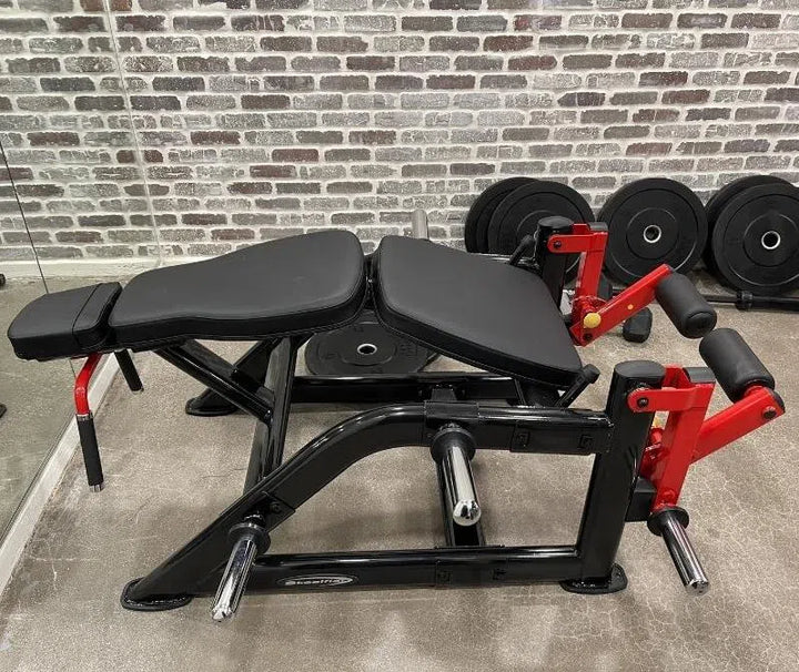 A Steelflex PLLC Hamstring Machine with black padding, red arm supports, and a sturdy black frame rests against a brick wall in the gym. Nearby, several black weight plates are neatly stacked on the floor, highlighting its commercial gym-quality equipment standards.