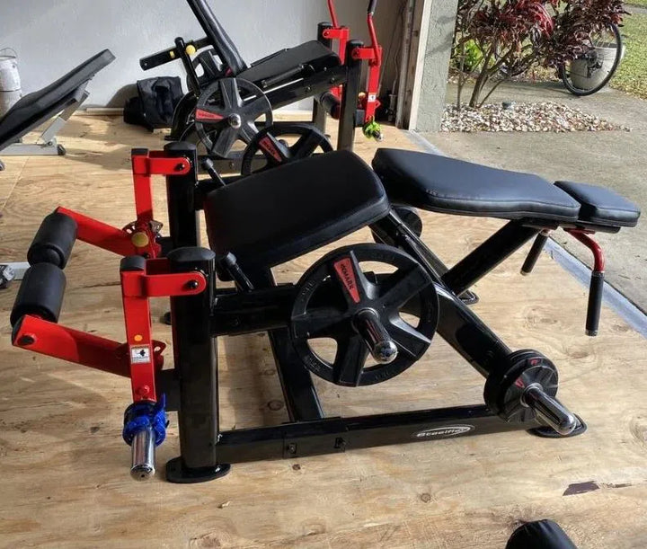 The Steelflex Hamstring Machine PLLC is showcased on a wooden floor, featuring red and black padding with adjustable settings. Weights are neatly attached, complemented by a bicycle and plants, highlighting its commercial gym-quality essence.