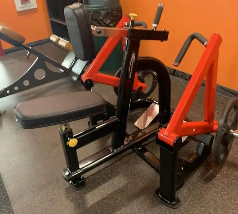 A red and black Steelflex PLSR rests on a rubber floor for seated exercises. It has an ergonomic design with padded seat, backrest with handles, and adjustable components against an orange wall. A commercial-grade workout bench is partially visible nearby.