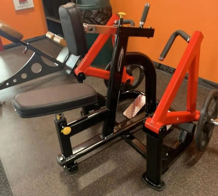 A red and black Steelflex PLSR rests on a rubber floor for seated exercises. It has an ergonomic design with padded seat, backrest with handles, and adjustable components against an orange wall. A commercial-grade workout bench is partially visible nearby.