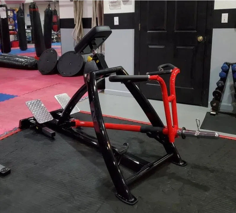The Steelflex T-bar Row Machine (PLTR), in black and red, is placed on a black mat. It boasts textured foot pedals and handles with red padding. This gym machine sits in a commercial gym room filled with mirrors and martial arts equipment.