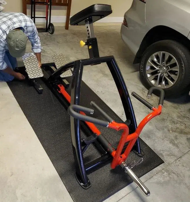In the garage beside a gray car, someone is setting up a red and black Steelflex T-bar Row Machine (PLTR) on a black mat. Kneeling to adjust the metal footplates, they are turning this space into a mini commercial gym.