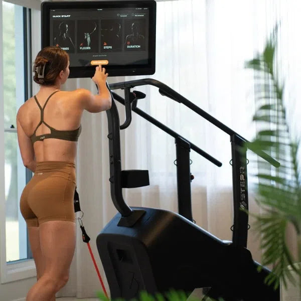 Wearing athletic gear, an individual uses a STEPR+ Stair Climbers touchscreen indoors. This space-saving home fitness machine shows workout stats such as steps and duration. Partial views of green plants in the foreground add a refreshing touch.