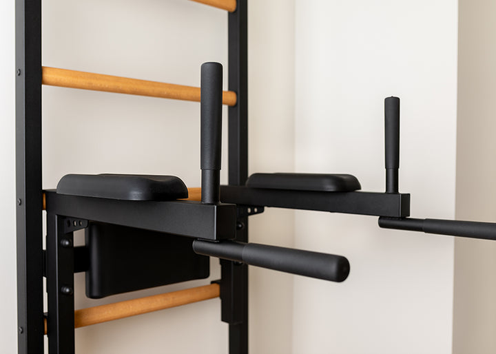 Close-up of the black BenchK Dip & Flex Pull-Up Bar on the BenchK Pro Gym Wall Bars 733, a wooden wall-mounted station with light-colored bars and padded supports for strength exercises.