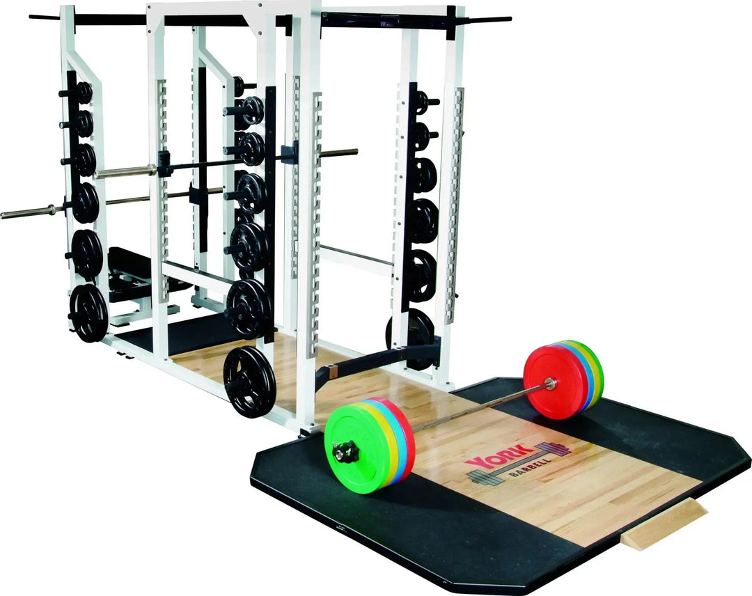 A York STS Triple Combo Power Rack holds a barbell with colorful plates in a gym setting, alongside extra weights. The wooden platform, branded York Barbell, complements the setup for strength training.