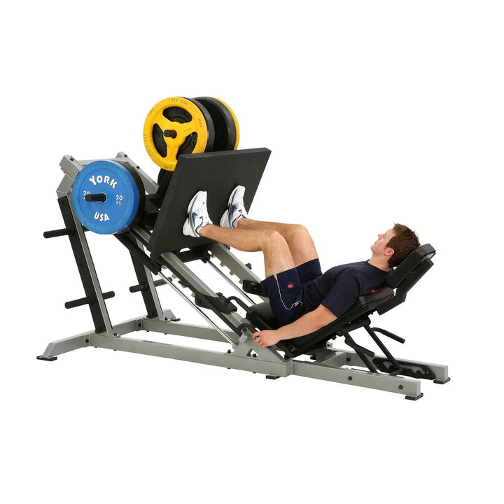 A person uses the York STS 35 Degree Leg Press Machine from York Barbell, pushing against a weighted platform with blue and yellow plates. Theyre wearing a navy shirt, shorts, and white sneakers while working out on the sleek silver and black equipment.