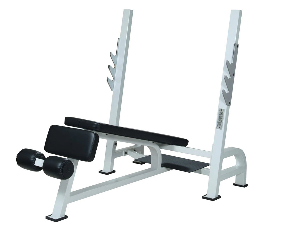 The York Barbell STS Olympic Decline Bench Press features an angled design, a white metal frame, and a black padded seat. It includes contour foam padding for comfort, curved leg support, and polished chrome bar holders with three hooks on each side.