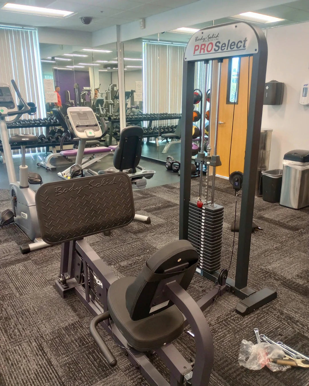 A gym with various exercise equipment, including a leg press machine labeled Pro Select and a stationary bike. The room has carpeted floors, large windows with blinds, and mirrors on the wall.